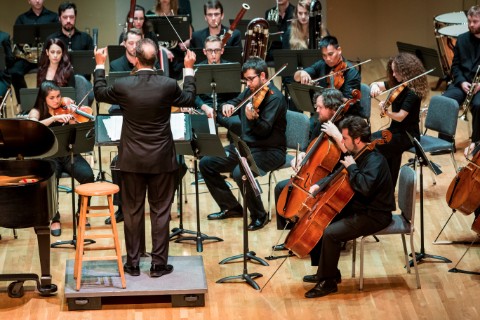 A conductor in a suit standing on a platform conducts an orchestra while they sit around him.